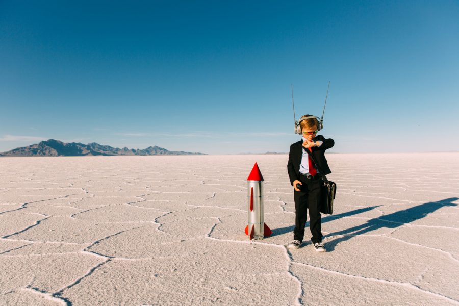 A young boy dressed in a business suit and tie stands on the Bonneville, Utah Salt Flats waiting for his new business and rocket to launch. He is wearing sunglasses and counting down to lift off while looking at his watch. The sky is blue and there is lots of copy space.